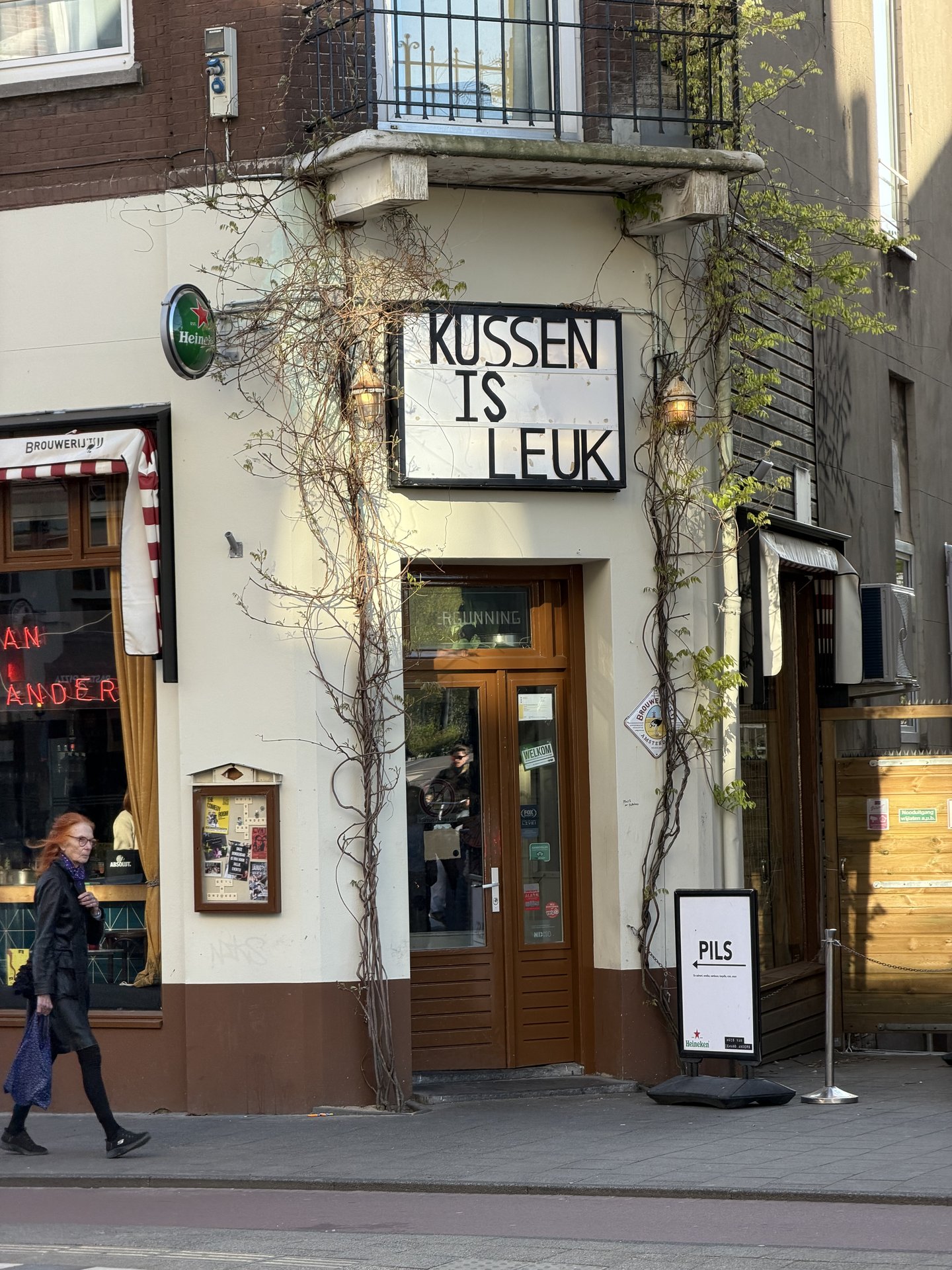 A person with red hair walks past a charming corner building on Stadhouderskade, Oude Pijp, Amsterdam, bathed in afternoon sun